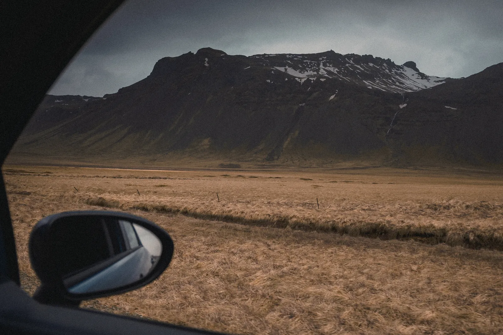 Snow-capped mountain range seen through a car's side window across dry grassland