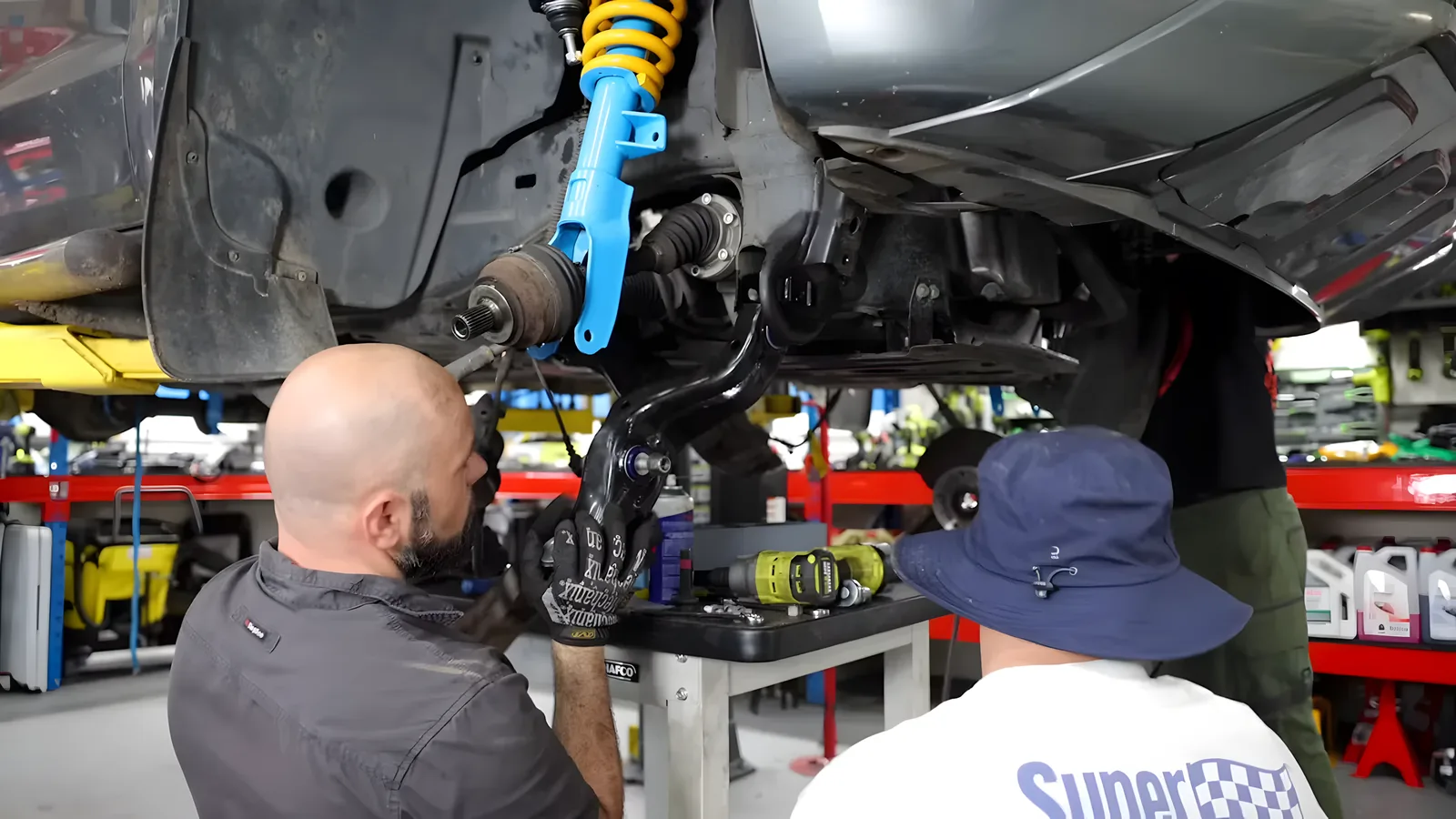 Two mechanics fitting a lower control arm and blue and yellow coilover strut to the front suspension of a car on a hoist